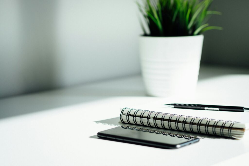 Minimalist desk setup featuring a spiral-bound notebook, a black pen, and a smartphone placed on a white surface. In the background, there is a white pot with a green plant, adding a touch of nature to the clean and modern workspace.