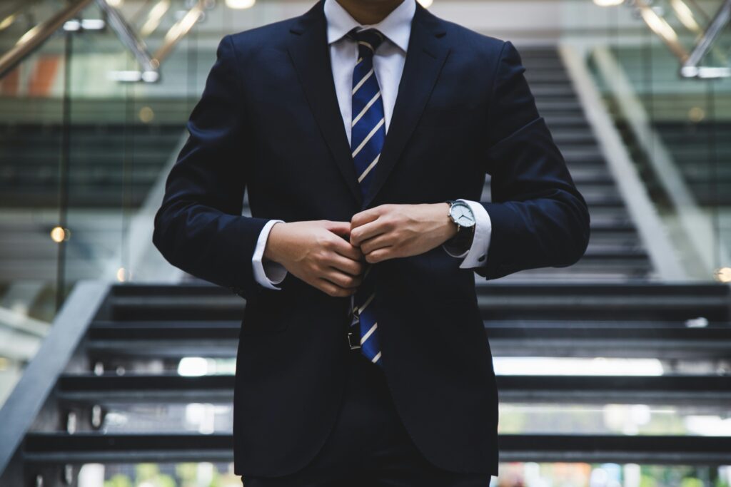 Business professional adjusting the button on a dark navy suit jacket while standing in front of a modern staircase. The individual is wearing a white dress shirt, a blue and white striped tie, and a silver wristwatch, conveying a polished, corporate appearance.}