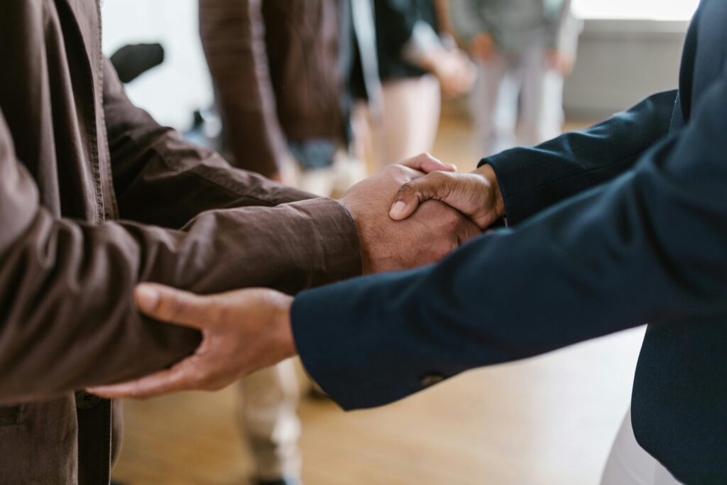 Two people shaking hands, both in long sleeve shirts—one navy blue and one brown—after discussing a business acquisition.