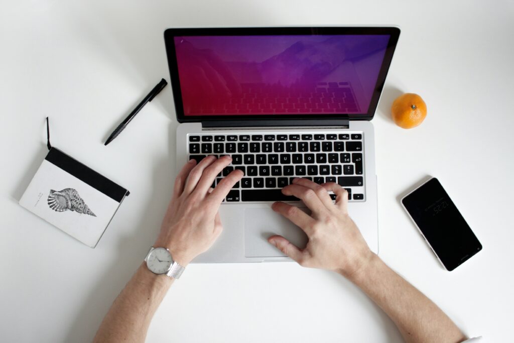 Image of a person's arms, typing on a laptop computer, researching the best software for tax practice management. On the white background, there is a notebook with a pen, along with an orange and a phone.