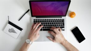 Image of a person's arms, typing on a laptop computer, researching the best software for tax practice management. On the white background, there is a notebook with a pen, along with an orange and a phone.