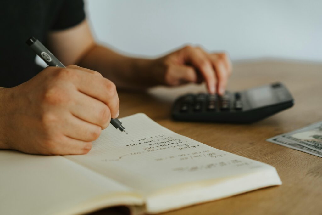 Close-up of a person writing calculations in a notebook with one hand while using a calculator with the other. A U.S. hundred-dollar bill lies nearby on the wooden table, suggesting a financial or budgeting task in progress.