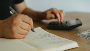 Close-up of a person writing calculations in a notebook with one hand while using a calculator with the other. A U.S. hundred-dollar bill lies nearby on the wooden table, suggesting a financial or budgeting task in progress.