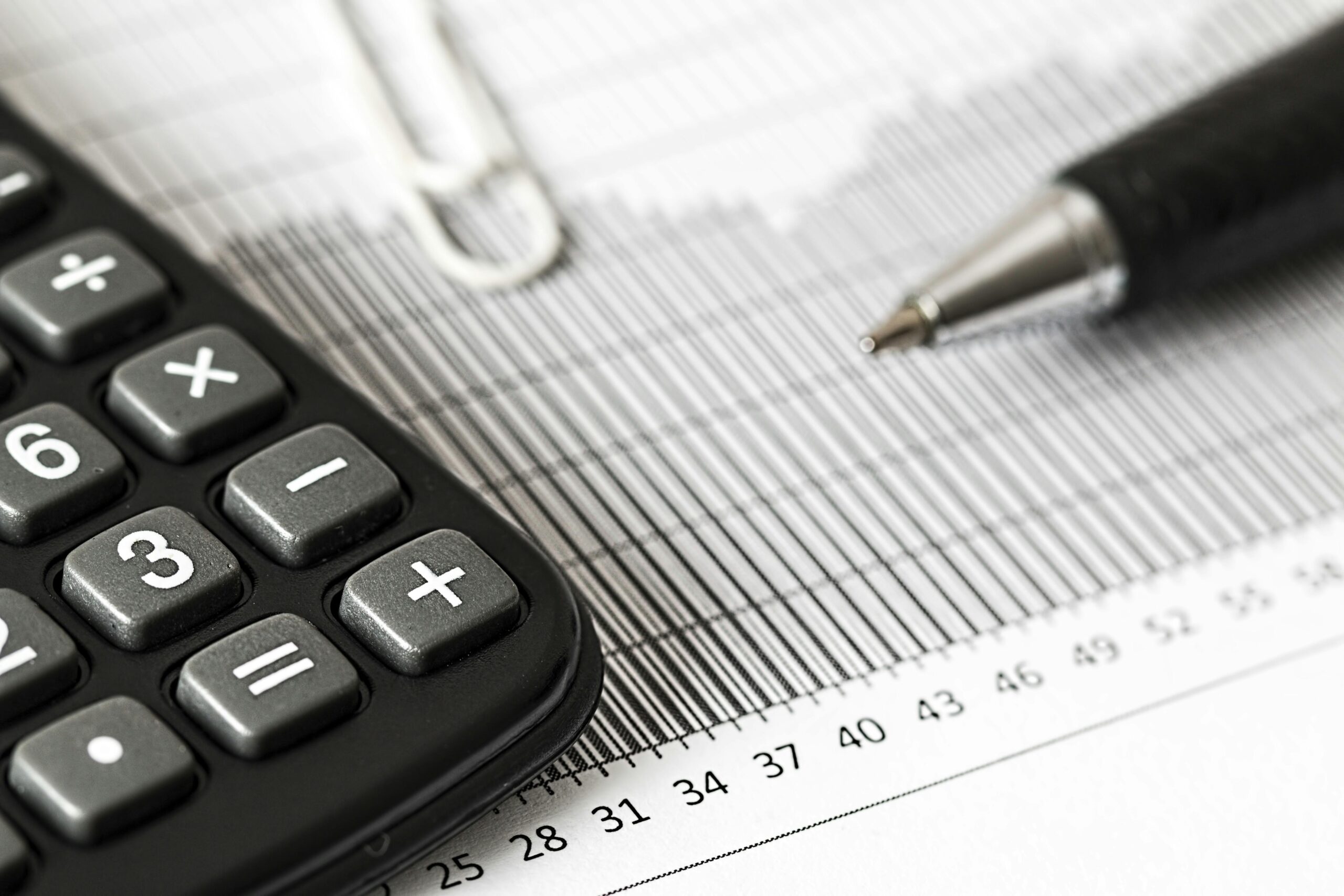 Close-up of a calculator, pen, and paperclip placed on a financial chart with horizontal and vertical lines. The image emphasizes financial analysis, accounting, or data calculations.