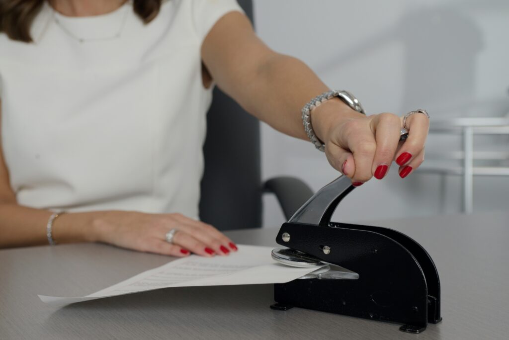 A woman in a white shirt writing on white paper with a pen, seated at a wooden desk. The scene conveys focus and professionalism in a business sale.