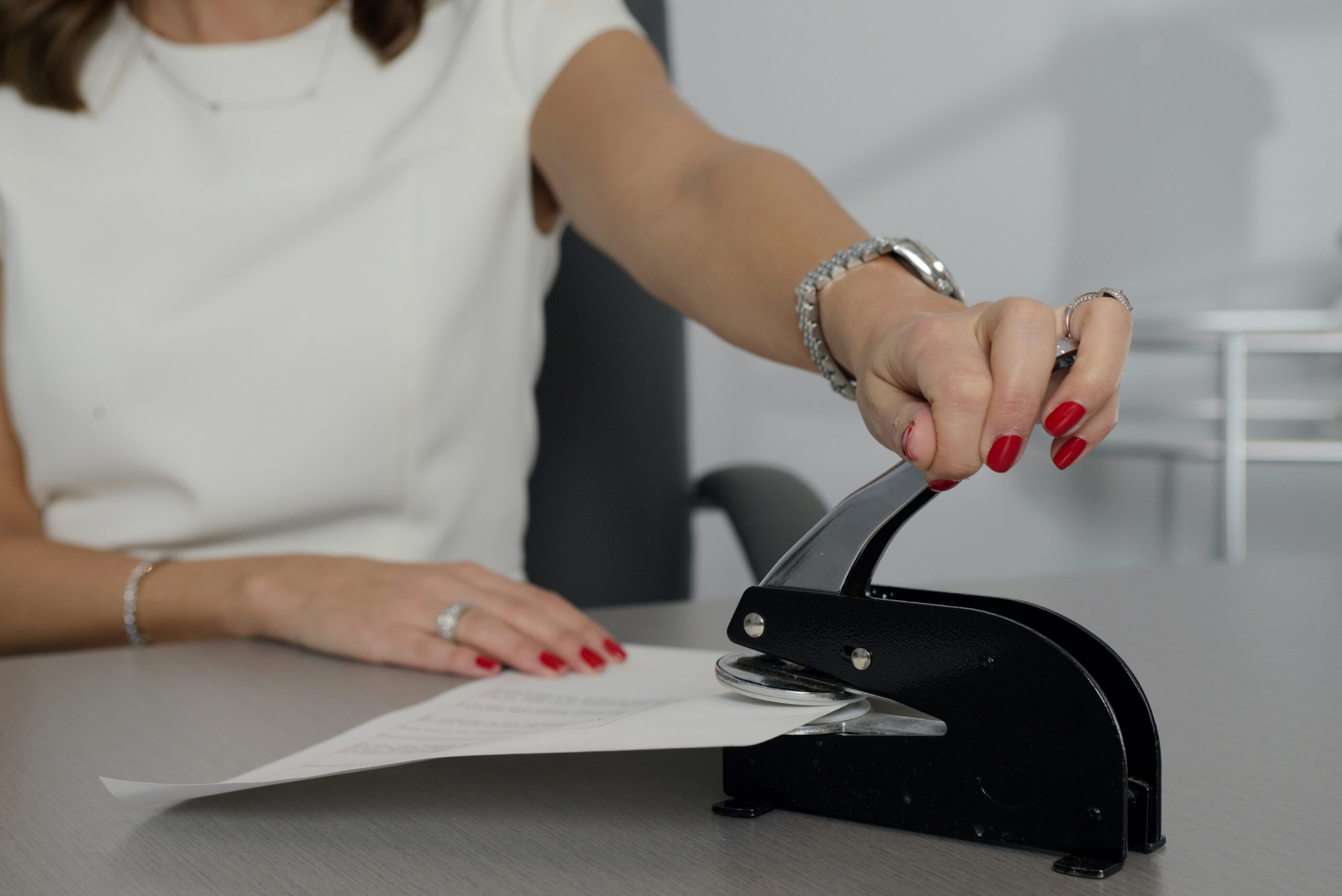 A woman in a white shirt writing on white paper with a pen, seated at a wooden desk. The scene conveys focus and professionalism in a business sale.