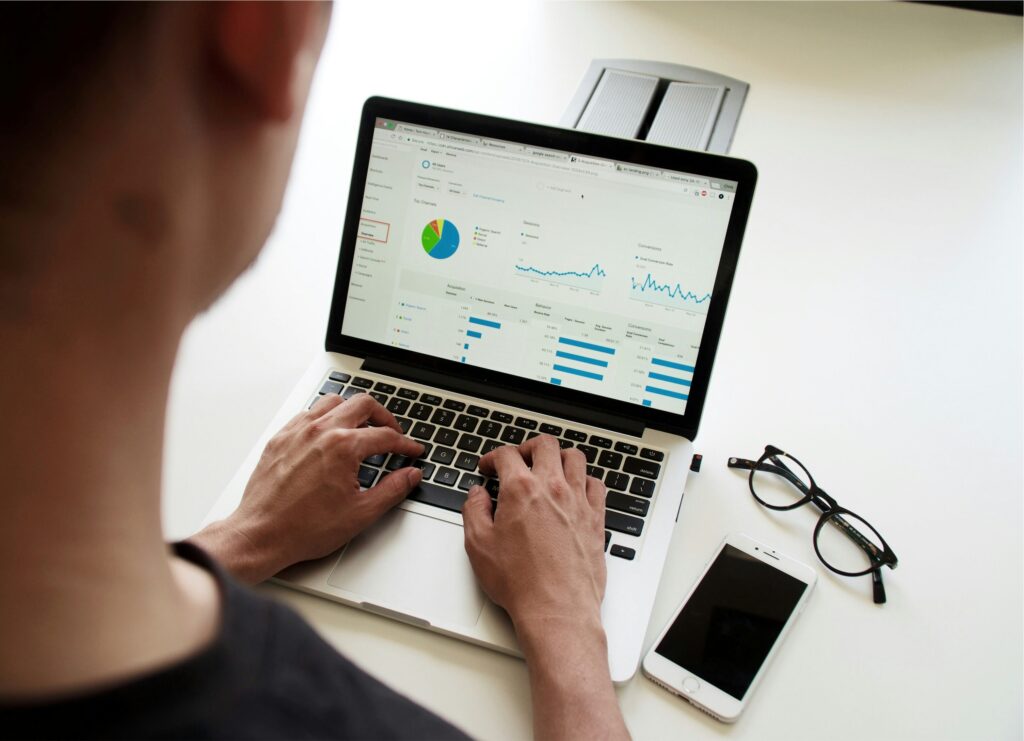 Person analyzing website analytics on a laptop, with various graphs, charts, and data visible on the screen. The individual is typing with both hands, and nearby on the white desk are a smartphone and a pair of black-rimmed glasses. The setting suggests a focus on digital marketing or data analysis.