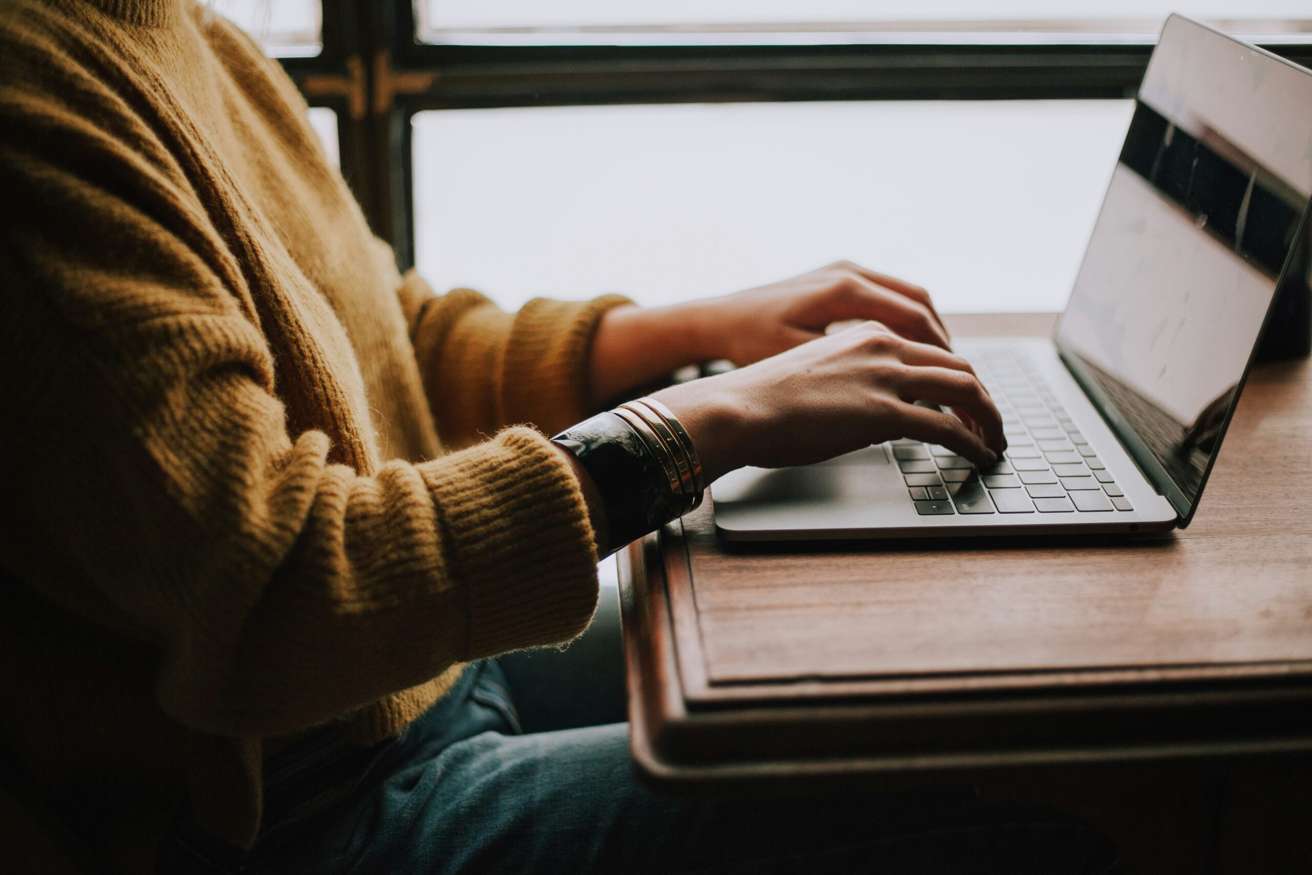 A person in a sweater wearing bracelets, typing on an open laptop, researching tax software options.