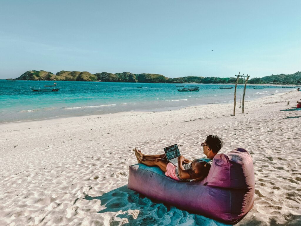 A man works remotely from a bean bag on the beach with a laptop, embodying the digital nomad lifestyle. The scene highlights the freedom and flexibility of location-independent work, along with the need to navigate cross-state or international tax considerations.