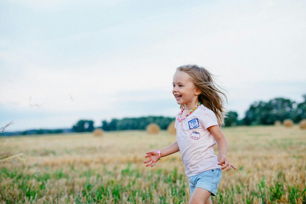 A young girl with long hair wearing a pink shirt, denim shorts, and colorful jewelry joyfully runs across a sunny field with hay bales scattered in the background. Trees line the horizon under a bright, open sky.