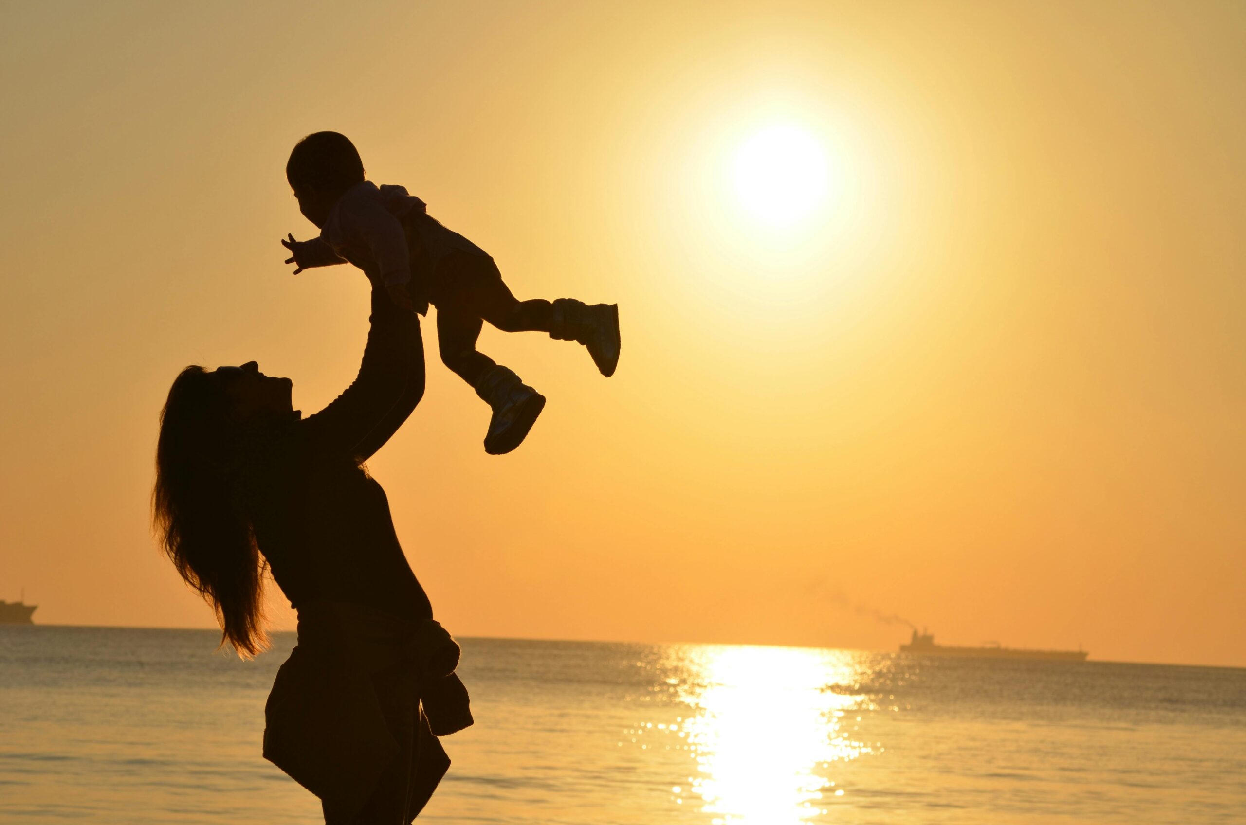 Silhouette of a mother joyfully lifting her child into the air at the beach during golden hour. The sun sets in the background, casting a warm, glowing light across the ocean and sky.