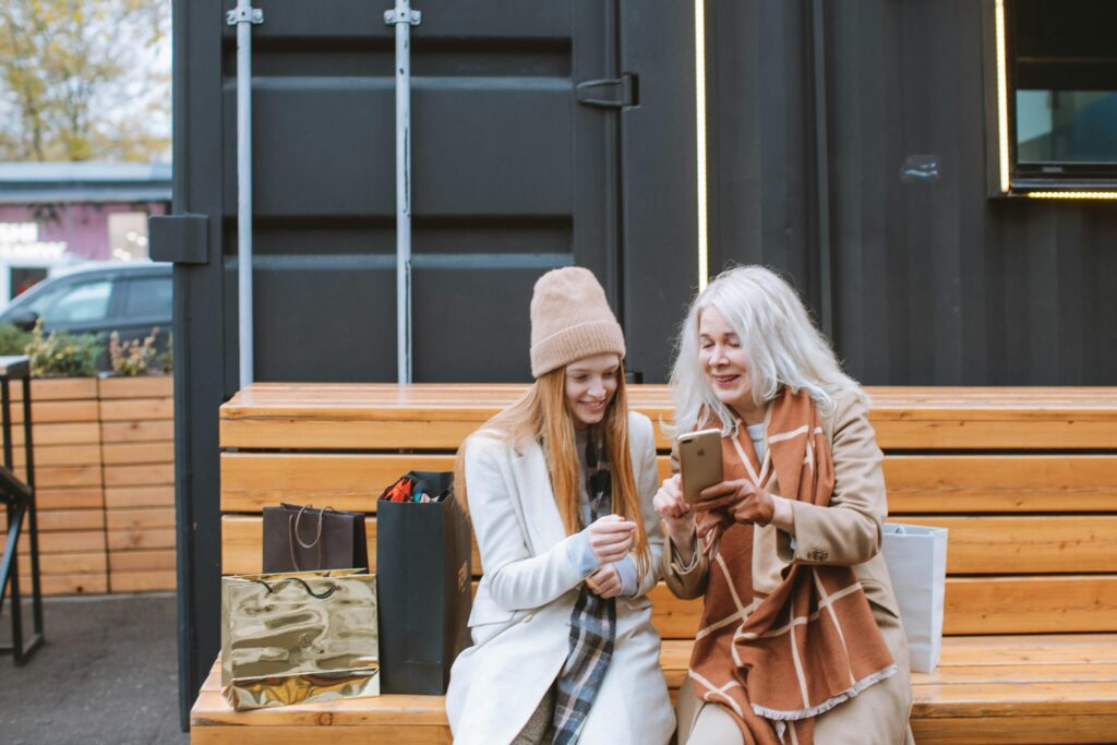 Retired woman sitting on a bench outdoors with her daughter while they both look at the older woman’s iPhone.