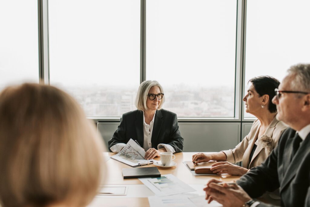 Silver-haired woman with glasses sits at the head of a full conference table in a meeting room with large windows.